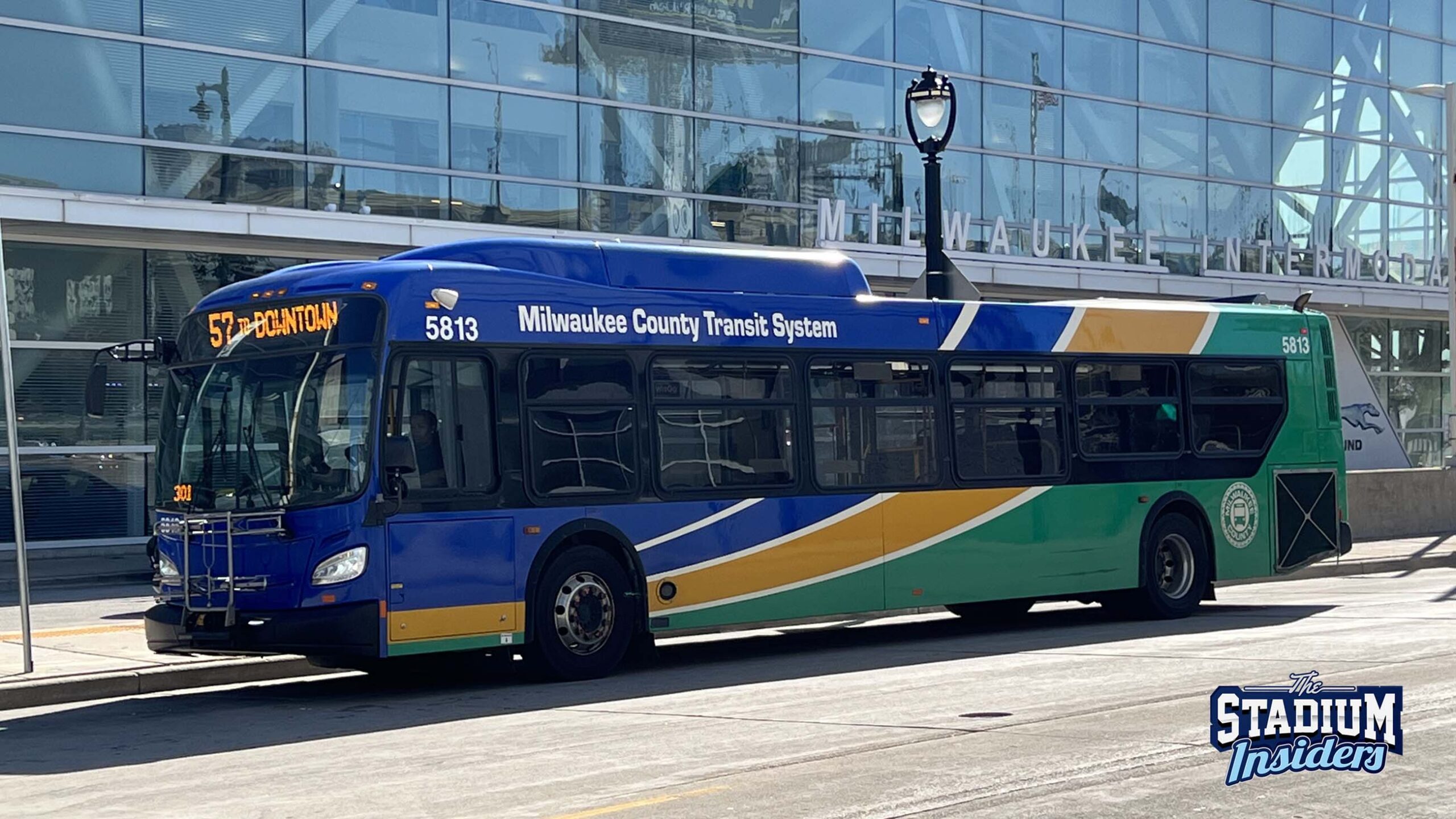 A blue yellow and green Milwaukee County Transit Bus is parked in front of the glass facade of the Intermodal Station